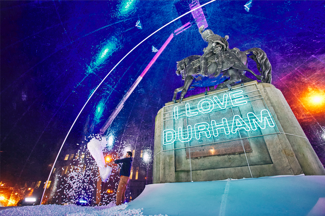 Man inside the snowglobe, empty a large bag with the fake snow onto the ground. Behind him is the statue of a man riding a horse on a plinth with the illuminated words I LOVE DURHAM on it.