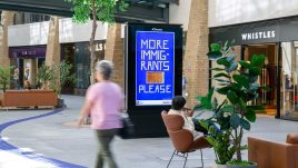 An image of a digital screen in a shopping centre showing the artwork ‘More Immigrants Please’ (2023) by Osman Yousefzada.