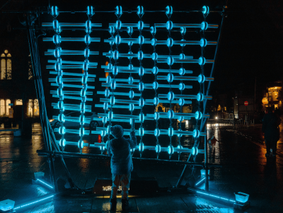 A light art installation seen at night. It's an interactive artwork, made up of a wall of glowing blue bars. A small child stands in front of it, engaging with the piece.