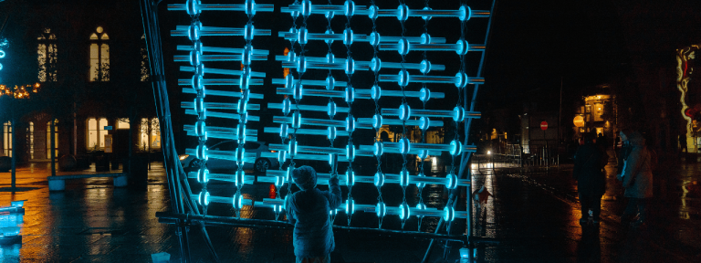 A light art installation seen at night. It's an interactive artwork, made up of a wall of glowing blue bars. A small child stands in front of it, engaging with the piece.