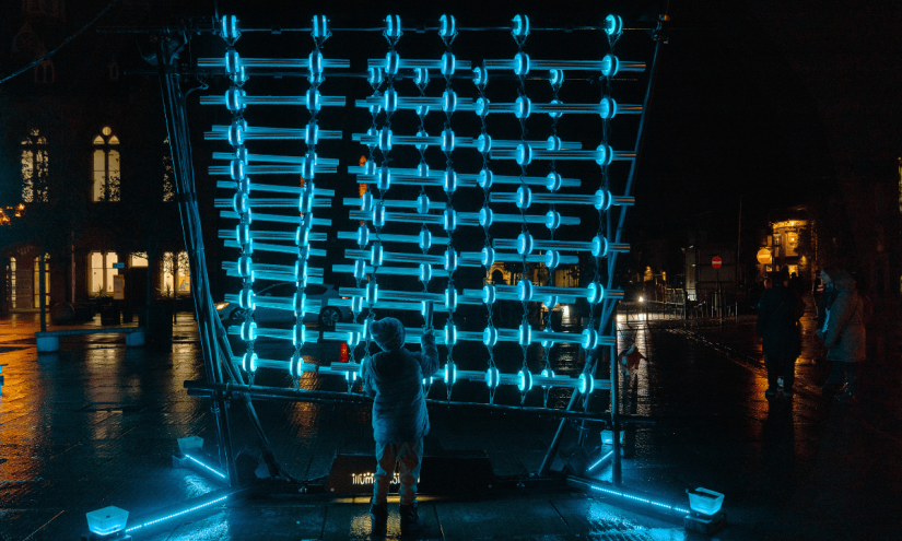 A light art installation seen at night. It's an interactive artwork, made up of a wall of glowing blue bars. A small child stands in front of it, engaging with the piece.