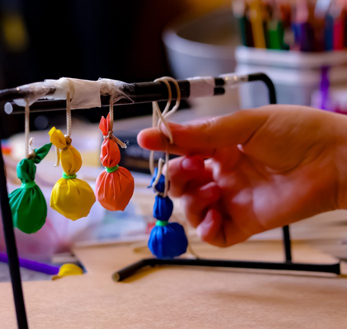 The image shows a close-up of a hand reaching towards a row of small colorful pouches tied with string, which are hanging from a horizontal rod. The pouches are of different colors—green, yellow, orange, and blue.