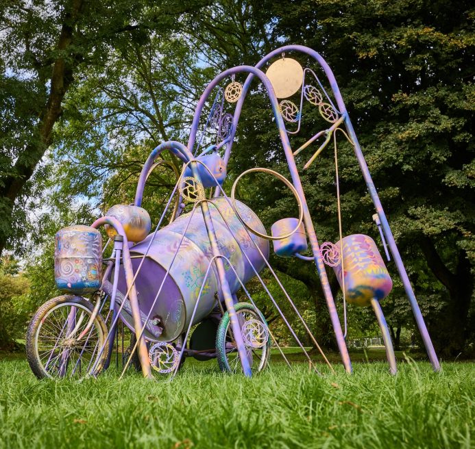 Race Leys Junior School and Spencer Jenkins' installation. The structure is comprised of large metal arches, bike parts and an old oil drum. The gears and steel drums have been spray painted blue, silver, purple, white, gold. Produced by Artichoke as part of Nuneaton & Bedworth Borough Council’s Creative Explorers programme. Photo by Andrew Moore