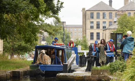 The photograph features a canal boat with passengers and what seems to be a sheep-like figure on board. People, some wearing life jackets, are gathered around the canal and engaging with the scene, suggesting an interactive or performative element to the event.