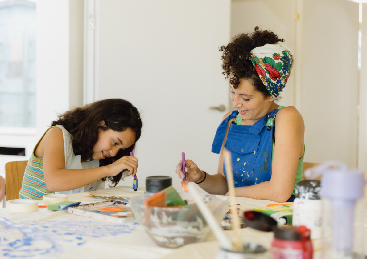 A young girl and a woman painting together. They are smiling and have paintbrushes in their hands. The table is covered in art materials.