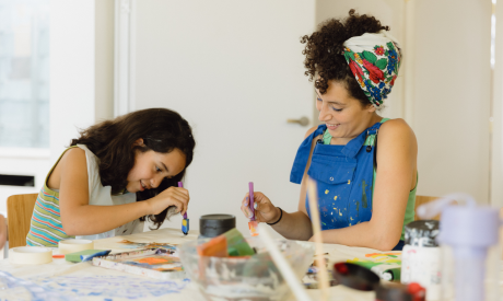 A young girl and a woman painting together. They are smiling and have paintbrushes in their hands. The table is covered in art materials.