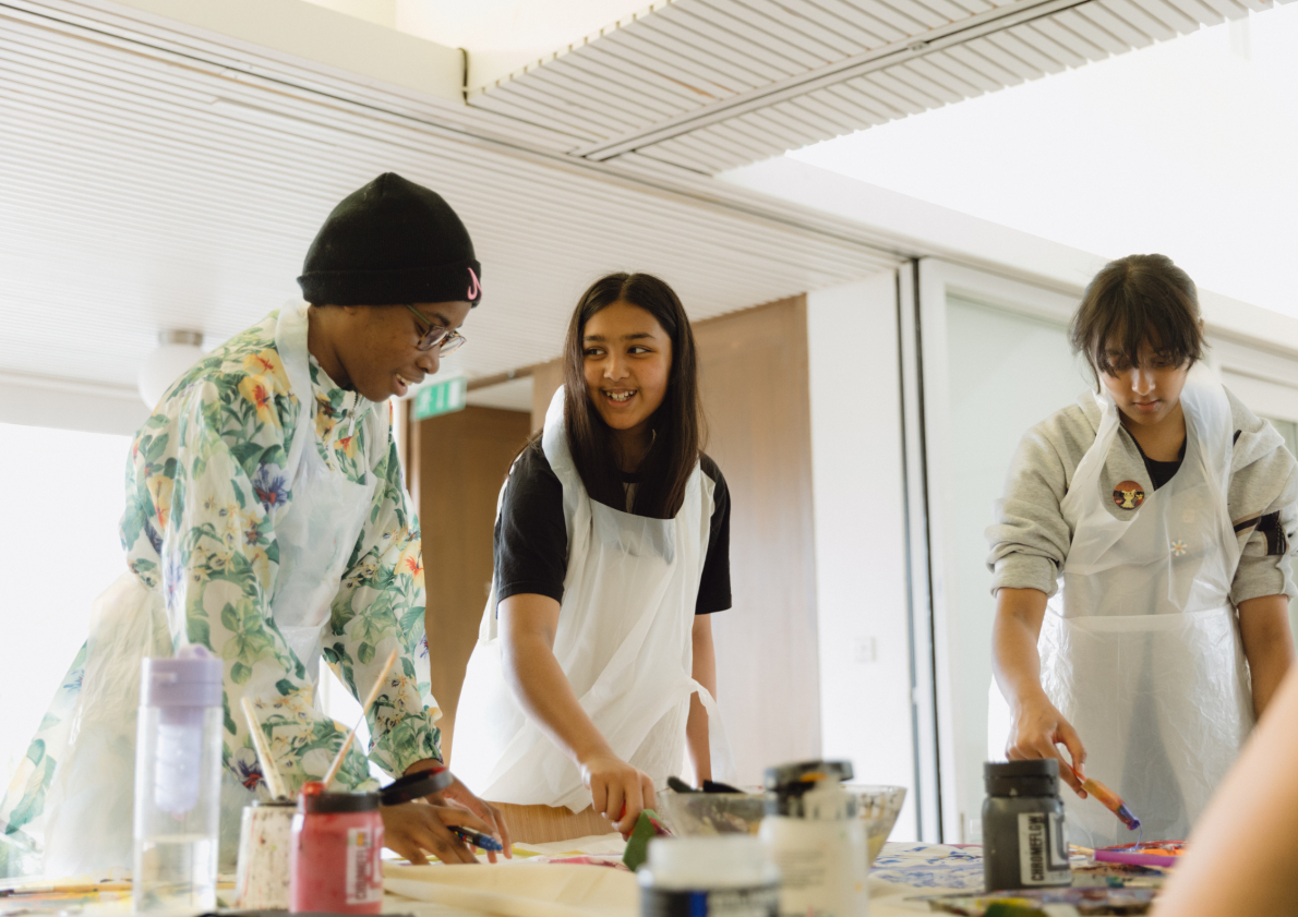 Three young girls wear aprons in a bright room. They are smiling and making art together,