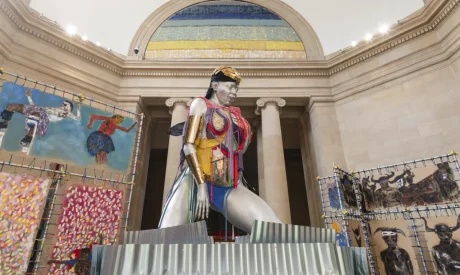 GRACE by Alvaro Barrington. The image shows a large, striking sculpture of a woman with metallic skin and colourful, mixed-media clothing, seated in a monumental pose within a classical building. Behind the figure are vibrant paintings featuring abstract human forms, adding to the dynamic atmosphere. The overall composition is bold, with bright colours, textures, and an imposing central figure.