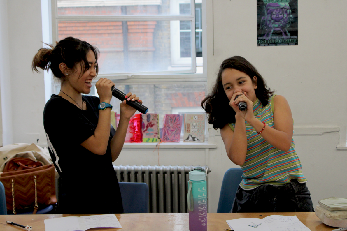 Two young participants share a joyful moment as they practice rap writing, holding microphones and smiling in a bright classroom. Creative artwork and personal items adorn the table and walls, adding to the vibrant atmosphere.