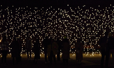 Light installation with hundreds of lightbulbs hanging down to create a rectangular shape. Within the shape the bulbs create you can see the figures of people standing