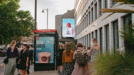 A busy city scene showing the artwork from The Gallery Season 4 on billboards. The artwork on display: A person with red hair stands confidently under the text 