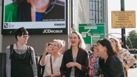 A city scene with the artists from The Gallery Season 4 looking at their art on billboards. The artwork on display: A man and woman are featured together in a split image. To the left is the womans face with lipstick and to the right is the man's with his glasses. The pair have contrasting hairstyles and makeup, illustrating their personal aesthetics.