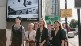 A city scene with the artists from The Gallery Season 4 looking at their art on billboards. The artwork on display: A black-and-white image showing legs sticking out of a kitchen cupboard.