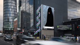 A busy city scene showing the artwork from The Gallery Season 4 on billboards. The artwork on display: A person wearing a black garment which appears to be a burqa with their face partially obscured. They hold a pearl in their mouth