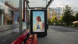 A busy city scene showing the artwork from The Gallery Season 4 on billboards. The artwork on display: A person with red hair stands confidently under the text 