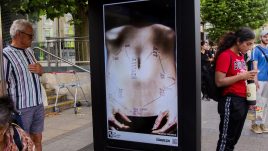 A busy city scene showing the artwork from The Gallery Season 4 on billboards. The artwork on display: A close-up of a person's bare midsection, with years written on their skin, connected by lines forming a path across the abdomen. The years range from 2014 to 2022, appearing multiple times in different locations. The person is lying down, with their hands resting on their lower abdomen, and a small tattoo is visible on their left arm. The image is set against a bed sheet.