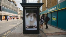 A busy city scene showing the artwork from The Gallery Season 4 on billboards. The artwork on display: A close-up of a person's bare midsection, with years written on their skin, connected by lines forming a path across the abdomen. The years range from 2014 to 2022, appearing multiple times in different locations. The person is lying down, with their hands resting on their lower abdomen, and a small tattoo is visible on their left arm. The image is set against a bed sheet.