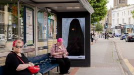 A busy city scene showing the artwork from The Gallery Season 4 on billboards. The artwork on display: A person wearing a black garment which appears to be a burqa with their face partially obscured. They hold a pearl in their mouth