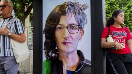 A busy city scene showing the artwork from The Gallery Season 4 on billboards. The artwork on display: A man and woman are featured together in a split image. To the left is the womans face with lipstick and to the right is the man's with his glasses. The pair have contrasting hairstyles and makeup, illustrating their personal aesthetics.