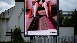 A busy city scene showing the artwork from The Gallery Season 4 on billboards. The artwork on display: A person wearing black lingerie and a cow mask sits in an armchair in a room with vintage wallpaper. They hold red feathered objects in each hand, with hairy legs spread wide. The image has a surreal, distorted effect, especially in the walls and floor, creating a warped perspective.