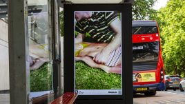 A busy city scene showing the artwork from The Gallery Season 4 on billboards. The artwork on display: A man wearing a striped shirt lies on the grass with their arm resting on a life-size cut out of a woman in swimwear.