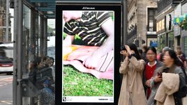 A busy city scene showing the artwork from The Gallery Season 4 on billboards. The artwork on display: A man wearing a striped shirt lies on the grass with their arm resting on a life-size cut out of a woman in swimwear.