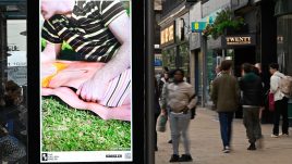 A busy city scene showing the artwork from The Gallery Season 4 on billboards. The artwork on display: A man wearing a striped shirt lies on the grass with their arm resting on a life-size cut out of a woman in swimwear.