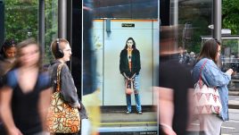 A busy city scene showing the artwork from The Gallery Season 4 on billboards. The artwork on display: A woman stands on a train platform wearing a fluffy black jacket and a bright orange bag. She looks uncomfortable