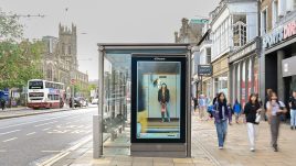 A busy city scene showing the artwork from The Gallery Season 4 on billboards. The artwork on display: A woman stands on a train platform wearing a fluffy black jacket and a bright orange bag. She looks uncomfortable