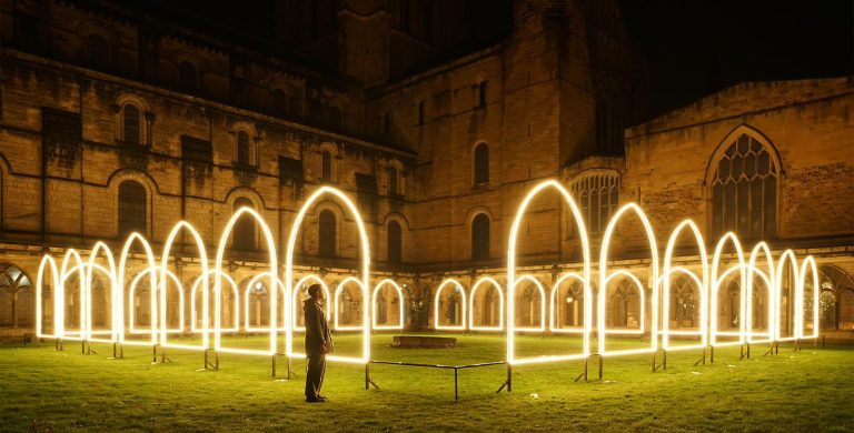 A person stands in front of a light art installation at night, looking at it in awe. The artwork is situated within a cathedral courtyard and the image is taken at night, making the artwork's glow even brighter. The installation features several frames that imitate the cathedral cloisters.