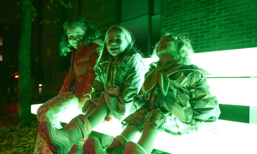 3 young girls sit together on a bench at night. The bench is glowing neon green. The girls are laughing.