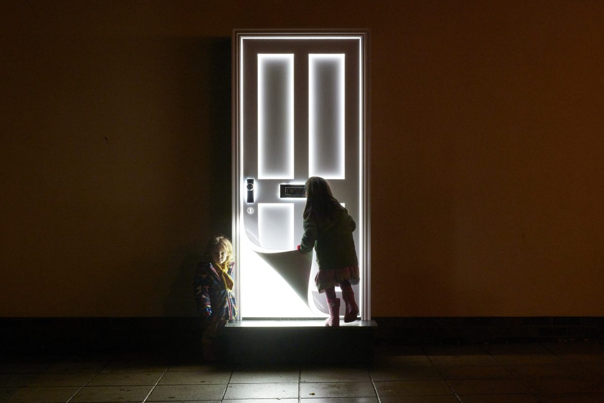 Two children stand before an illuminated door. The bottom left corner of the door seems to be peeling up like a piece of paper, revealing bright white light. the image is taken at night. The children look curiously at the artwork.