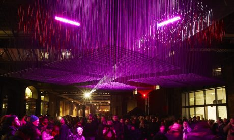 A crowd of people at night in Kings Cross London. They stand underneath an angular cluster of hanging lights. The sculpture and audience are bathed in purple and red light