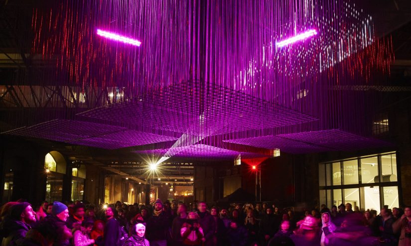 A crowd of people at night in Kings Cross London. They stand underneath an angular cluster of hanging lights. The sculpture and audience are bathed in purple and red light