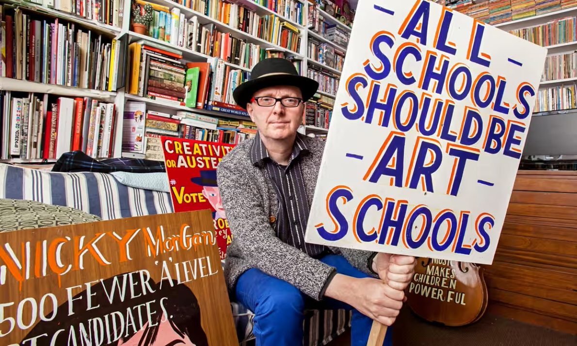 A man wearing a hat and glasses sits in a room filled with books, holding a large protest sign that reads, 